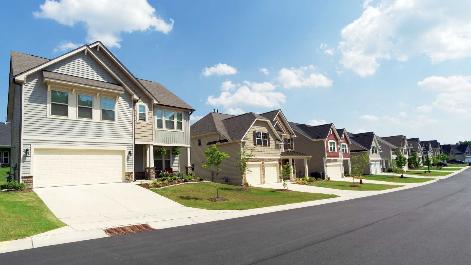 A suburban street lined with modern houses featuring landscaped yards under a blue sky with fluffy clouds.