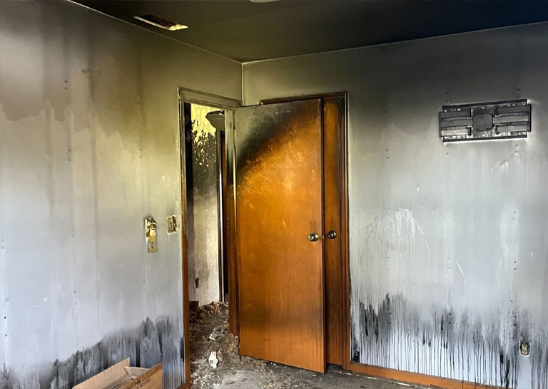 room-charred-walls A room with charred walls and a blackened door, indicating significant fire damage, with soot stains and debris on the floor.